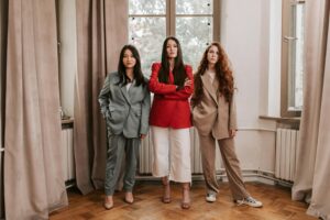 why some people look more attractive even without makeup Three women confidently pose indoors, showcasing stylish business attire against elegant curtains.