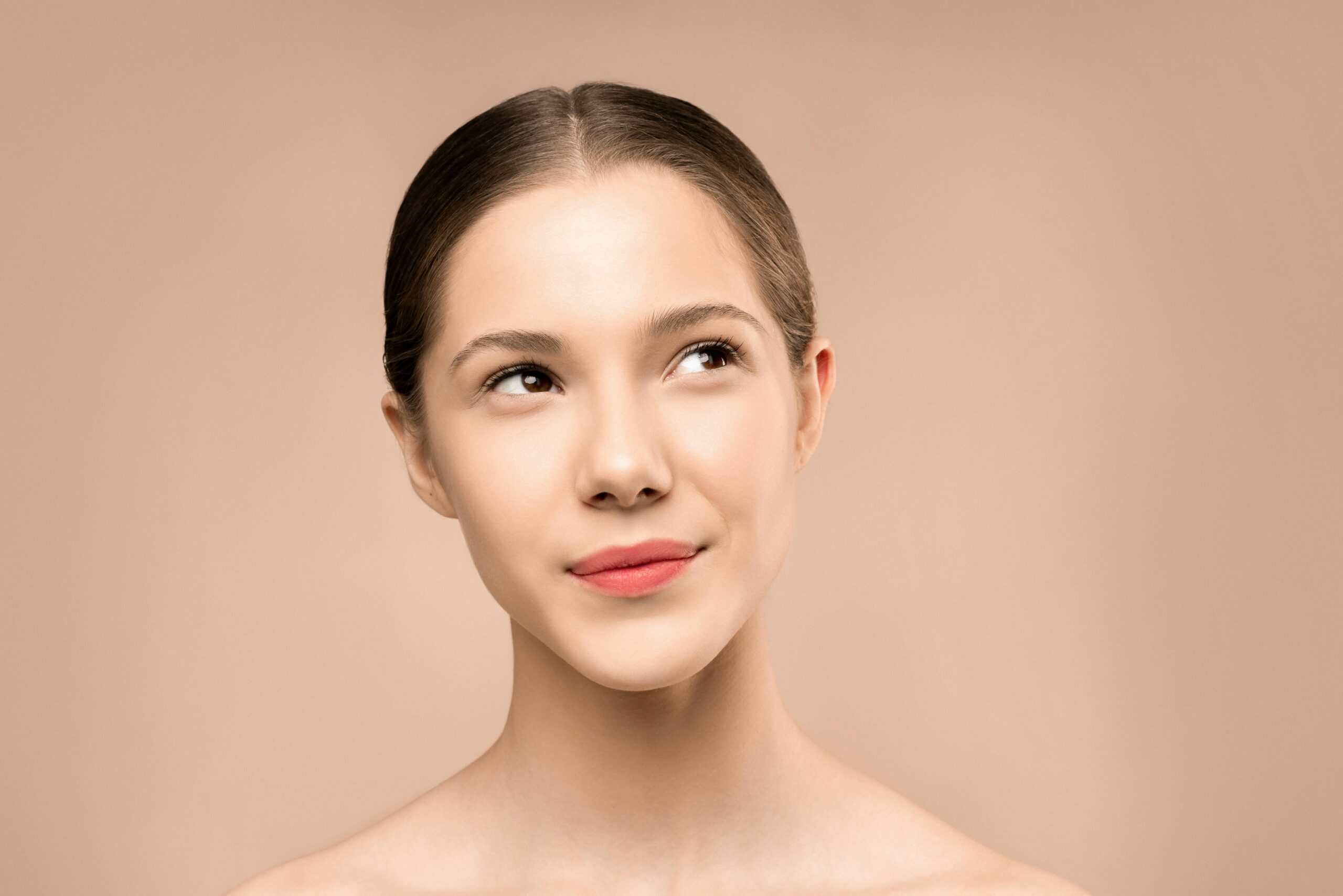 A young woman with radiant skin looks upward against a neutral background, embodying natural beauty.